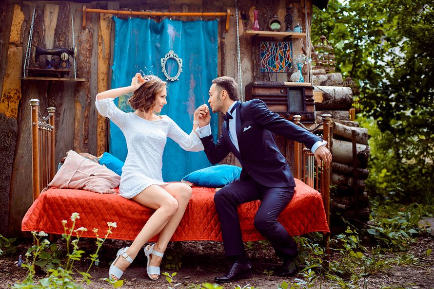 Возле деревянного домика с голубой шторкой, на красной кровати с голубыми подушками сидят жених и невеста - фото 2651009 Фотограф Владислав Озеров
