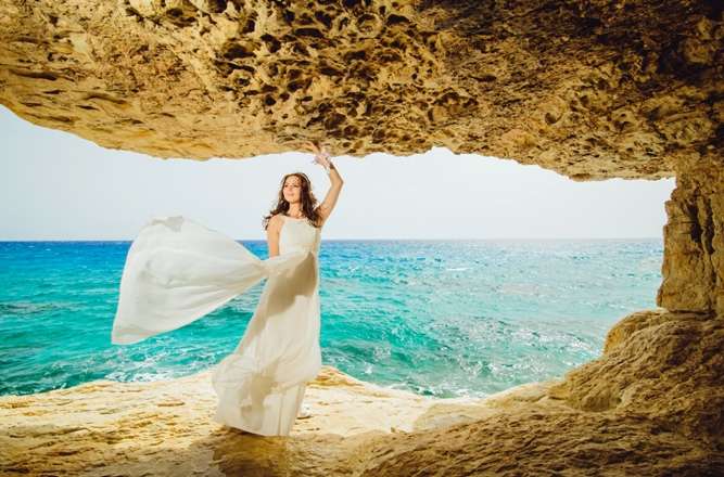 В проеме пещеры на фоне лазурного океана стоит невеста в белом платье, шлейф которого развивается на ветру,  поддерживая рукой - фото 3089681 Агентство Гименей - организация свадеб на Кипре