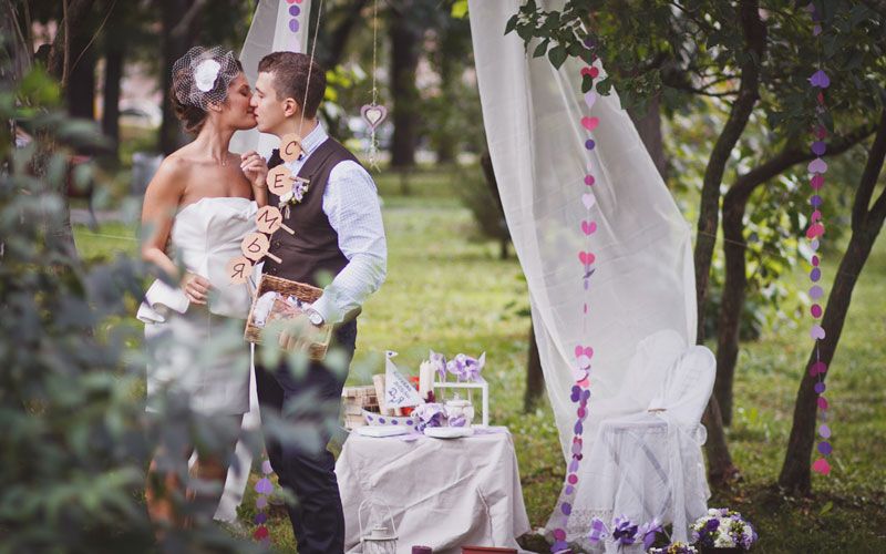 Жених и невеста стоят возле белого занавеса на фоне деревьев, украшенных гирляндами и фотографиями - фото 1076267 Фотограф Ведута Валерий