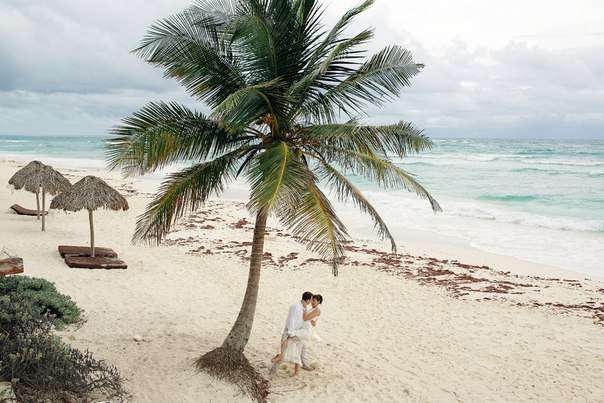 Возле пальмы на белом песке возле моря, молодожены целуют друг друга, в белых одеждах, на пляже стоят бамбуковые зонты - фото 2284878 Travel Service Mexico - свадьба в Мексике