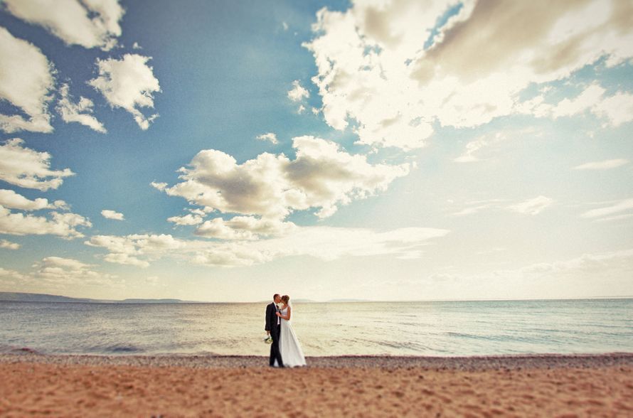 На пляже стоят жених и невеста, обнимая друг друга, она в белом платье, он в черном костюме - фото 1529747 Фотограф Александр Климов