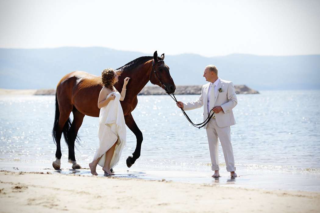 На пляже, жених держит лошадь, в белом костюме, рядом стоит невеста, в белом, длинном платье - фото 1529755 Фотограф Александр Климов