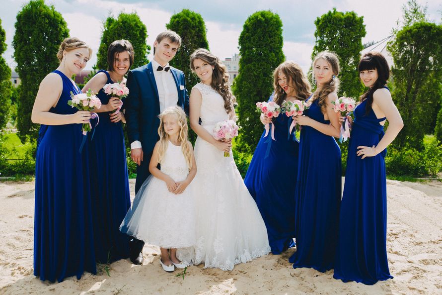 Жених в синем костюме, бабочке обнимает невесту в платье А-образного силуэта рядом девочка в белом платье "принцесса" и подружки в одинаковых синих длинных платьях держат букеты на пляже - фото 3174267 Nevesta AngelinA