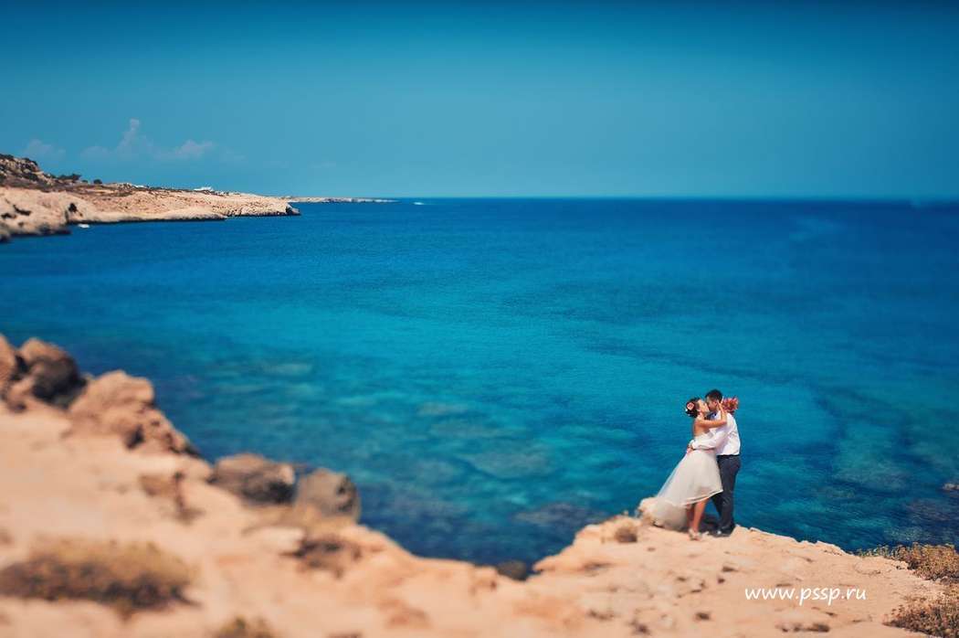 На пляже стоят на скале, обнявшись жених и невеста, она в белом, коротком платье, со шлейфом, он в белой рубашке и черных брюках - фото 1635047 Olya_shastie