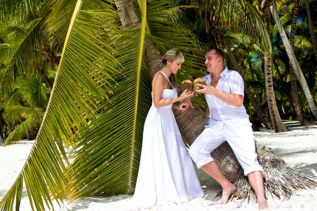 Жених стоит возле пальмы, в руке держит  фрукт, в голубой рубашке и белых шортах, невеста стоит в белом длинном платье со шлейфом, - фото 1942351 Okko group - организация свадьбы в Доминикане