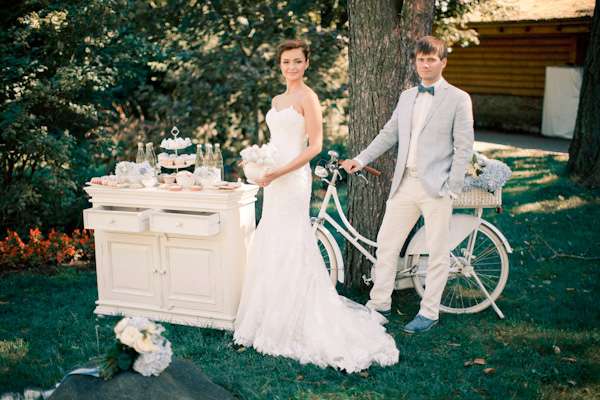 Жених и невеста стоят на фоне белого велосипеда, зеленого дерева возле бежевого комода - фото 1714577 Студия свадеб Oh Marriage
