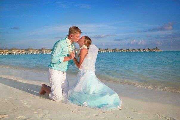 На песчаном пляже, стоя на коленях, целуются влюбленные, жених в голубой рубашке, невеста в голубом платье, на шее у них ожерелья - фото 2572501 Агентство Only event - организация свадьбы