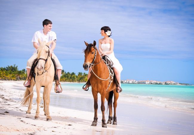 На пляже, жених сидит на белой лошади в светлом костюме, невеста сидит на коричневой лошади, в белом коротком платье, улыбаясь - фото 1915961 Tez Tour