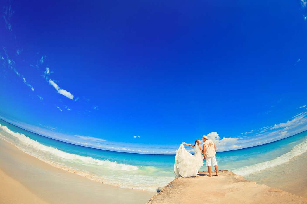 На каменистом пирсе парень в белой безрукавке, шортах и шляпе держит за руку невесту в белоснежном пышном платье, она его держит - фото 2606101 Dave Primov - protographer
