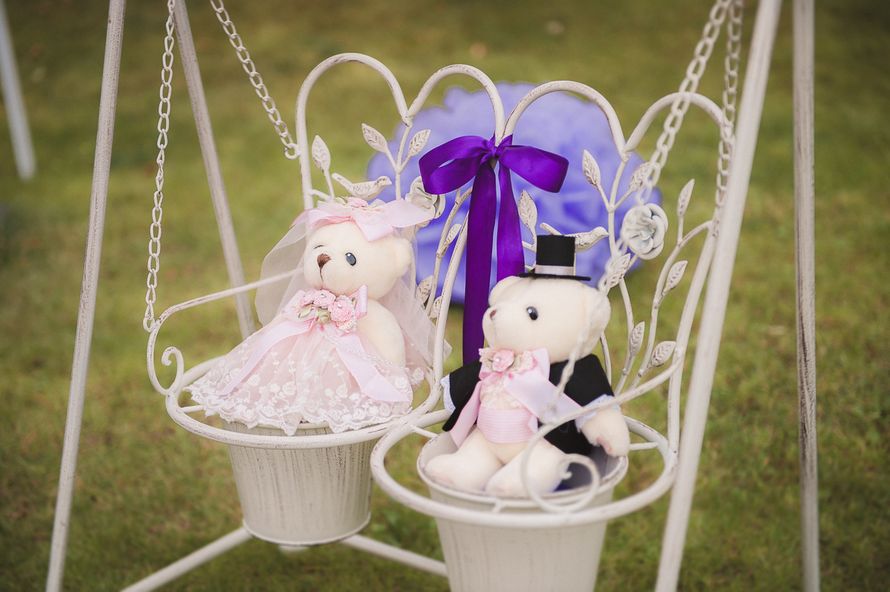 На игрушечных белых качелях, украшенных фиолетовым бантом, сидят плюшевые медвежата - фото 2992077 Fleren