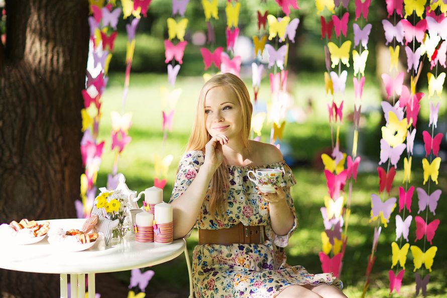 За маленьким белым столиком, между гирлянд из розовых, жёлтых и сиреневых бабочек сидит в цветочном платье девушка - фото 2625799 Флориника - студия флористики и декора