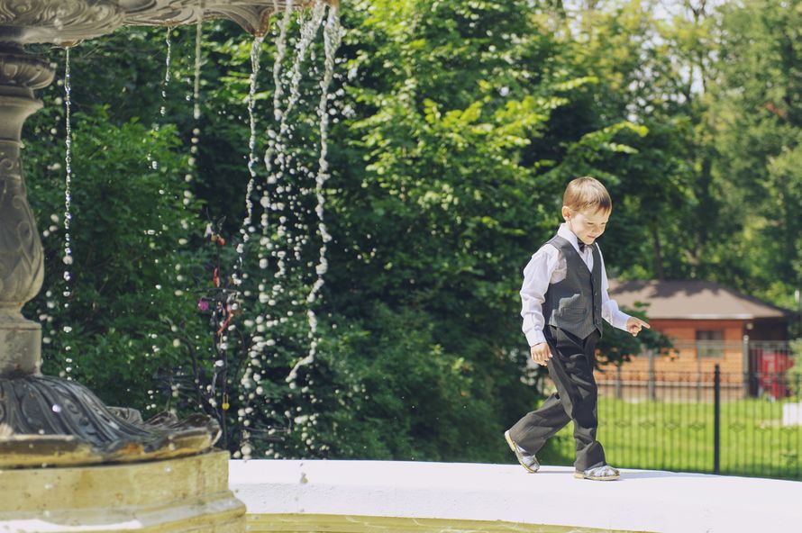 Мальчик в летний солнечный день на свадьбе бежит по фонтану в белой рубашке сбабочкой в серой жилетке с черными брюками - фото 2646699 Фотограф Мария Алексеева