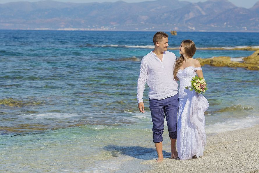 По кромке океана гуляют влюбленные, любуясь друг другом, жених в синих брюках и белой рубашке держит за талию невесту в белом - фото 2766609 "Tytprod" - фото и видеосъёмка 