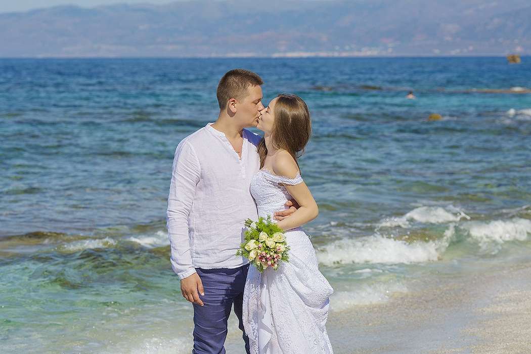 На тропическом пляже целуются молодожены, жених в синих брюках и белой рубашке держит за талию невесту в белом платье, она - фото 2766613 "Tytprod" - фото и видеосъёмка 