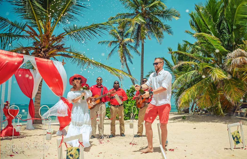 На тропическом побережье на фоне свадебного шатра в бело-красных тонах жених в коралловых бриджах и белой рубашке открывает - фото 2858239 Soul X-Ray Photo and Video в Доминикане