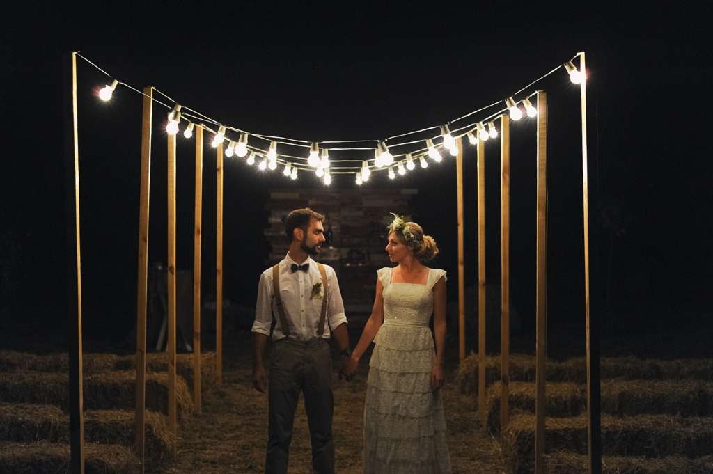 Жених и невеста стоят, держась за руки возле стогов сена под гирляндами из лампочек - фото 3144913 Svetlja4ok