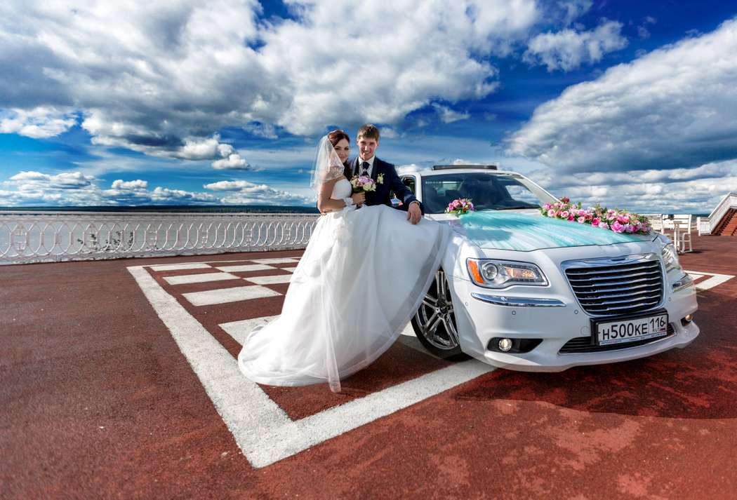 Белый свадебный "Chrysler" украшенный свадебной атрибутикой на фоне молодоженов - фото 3248101 Фотограф Александр Тилинин