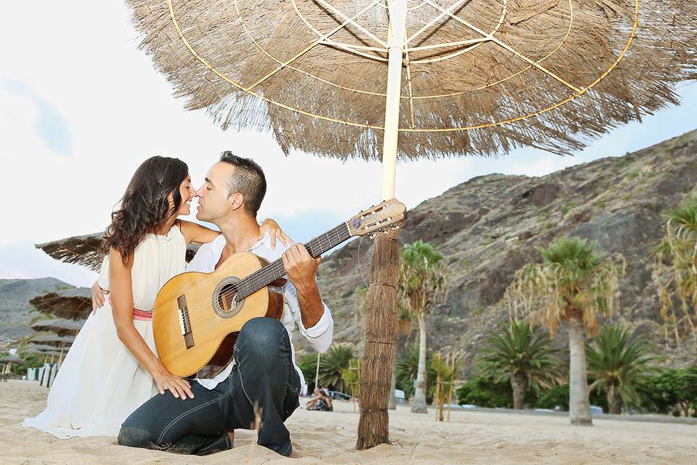 Под пляжным зонтиком сидят молодожены, жених в темных брюках и белой рубашке держит в руках  гитару и смотрит на невесту в белом - фото 3388819 BestDayTenerife - свадьба на Тенерифе