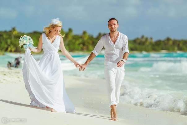 По пляжу идут жених и невеста, взявшись за руки, она в белом, длинном платье, с букетом цветов, он в белой рубашке и брюках - фото 3566357 Полина1303