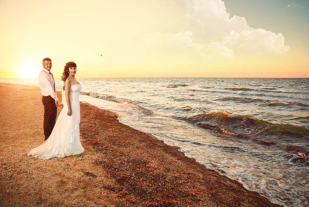 На пляже стоят жених и невеста взявшись за руки, она в открытом, кружевном платье, со шлейфом, он в белой рубашке и черных брюках - фото 3668055 Арт фотограф Ренат Хисматулин 