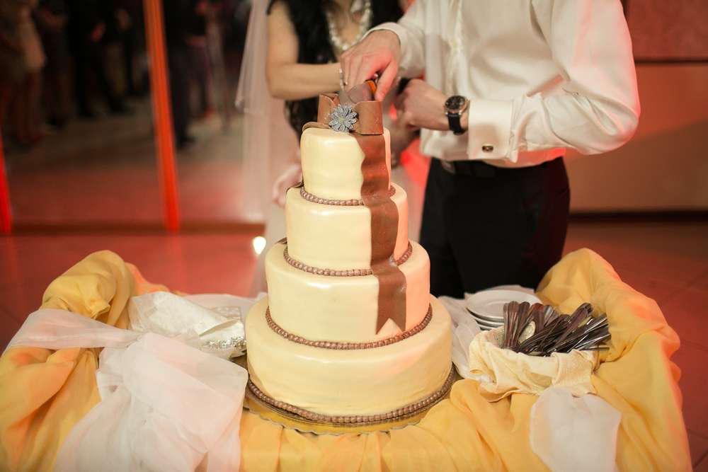 Жених с невестой, разрезающие белый 
свадебный торт, украшенный коричневым бантиком из мастики  - фото 934209 Фотограф Alex Paul