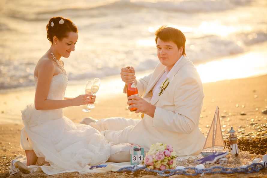 На песчаном пляже отдыхают молодожены, жених в белом костюме открывает шампанское,  невеста в открытом белом платье держит в - фото 614862 webgirl99