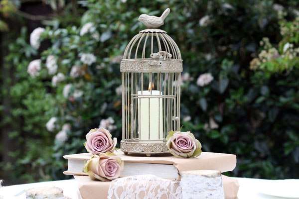 На фоне зелени на столе стоят стопка книг, розовые цветы, клетка со свечкой - фото 953225 Салон свадебной моды "Карамель"
