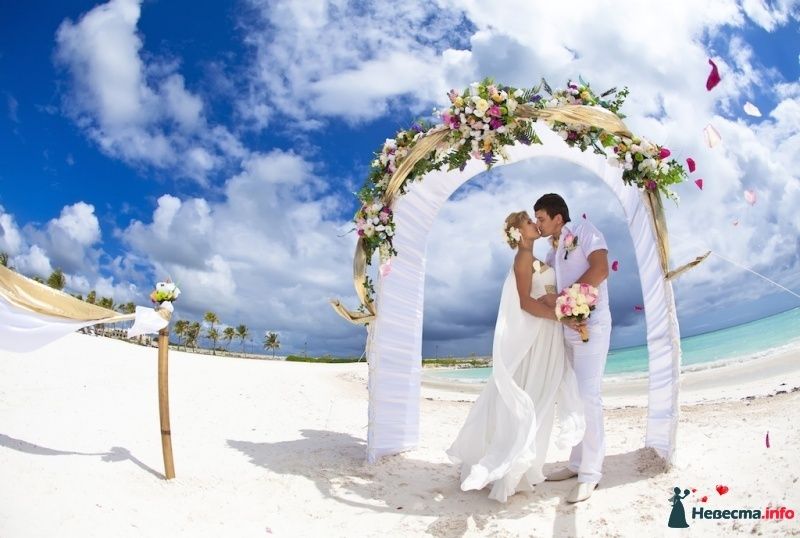 Под сводом свадебной арки, украшенной белой тканью и цветами, стоят молодожены, слившись в поцелуе, жених в белых брюках и - фото 452699 Caribbean Wedding - свадьба в Доминикане