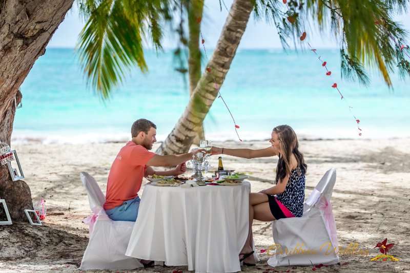 На тропическом пляже в тени деревьев за столиком, застеленным белой скатертью, сидит влюбленная пара, девушка в синем платье, - фото 2810969 Caribbean Wedding - свадьба в Доминикане